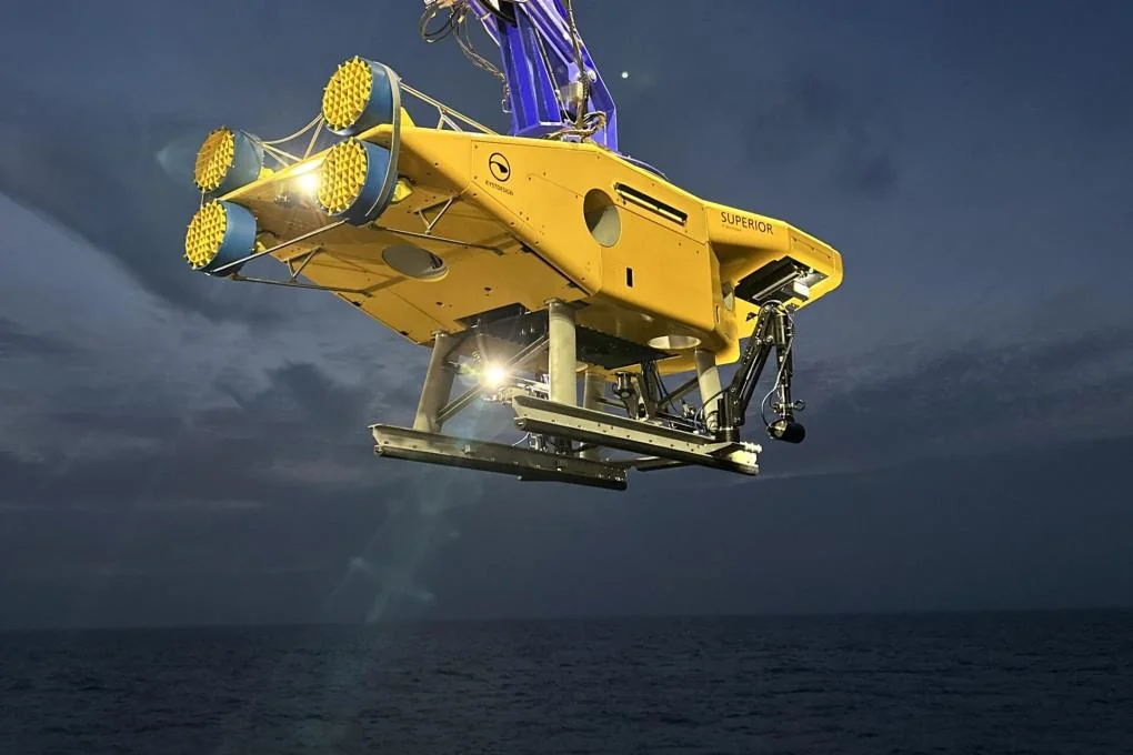 Veicolo subacqueo giallo sospeso sopra il mare con telaio inferiore e cavi di sollevamento.